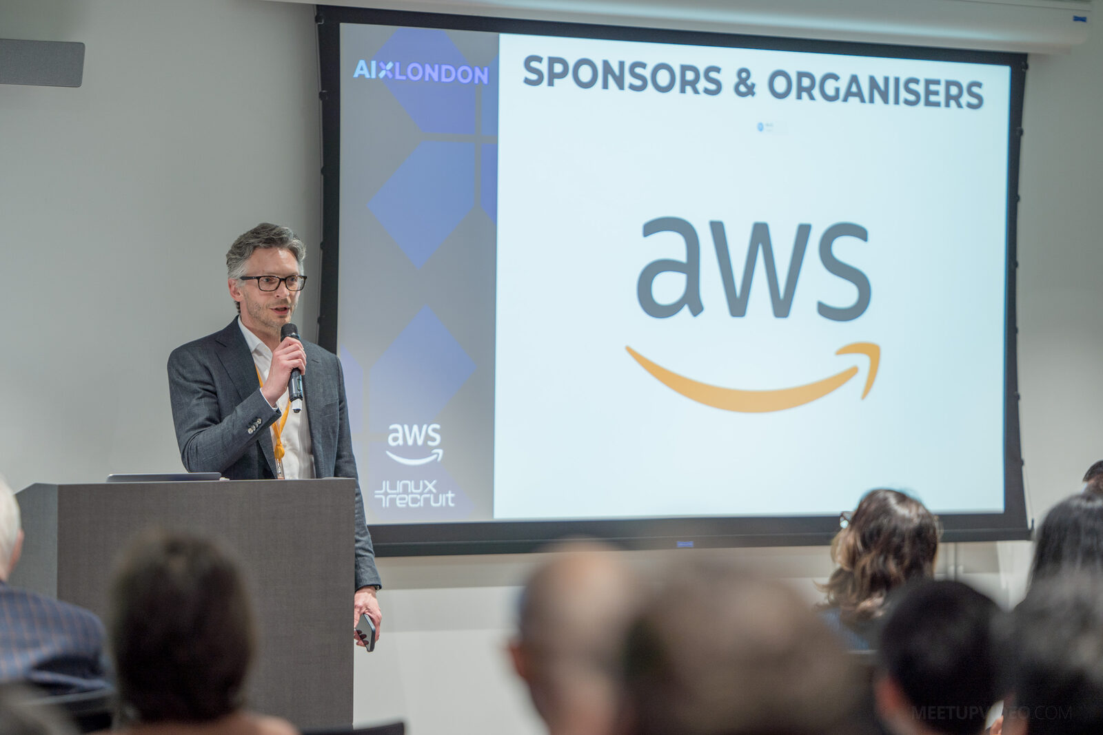 A host opening AI Exchange @ AWS at the lectern, microphone in hand, beside a screen showing the AWS logo.