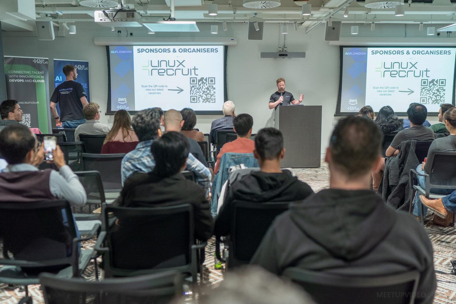 A full room seen from the back, a speaker at the lectern and twin screens showing the Linux Recruit sponsors slide with a QR code for current vacancies.