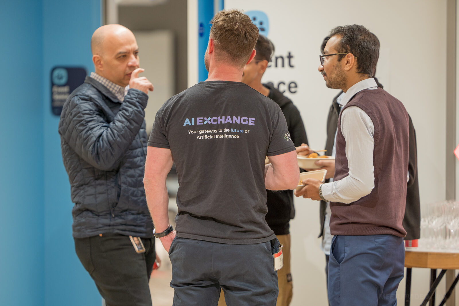 Three attendees talking over food before the talks, one of them wearing an AI Exchange t-shirt reading “your gateway to the future of Artificial Intelligence”.
