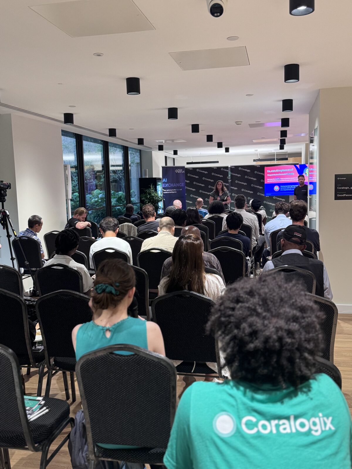 The audience seen from the back of the room, rows of seated attendees facing a speaker and a slide asking “So, can AI replace me?”.