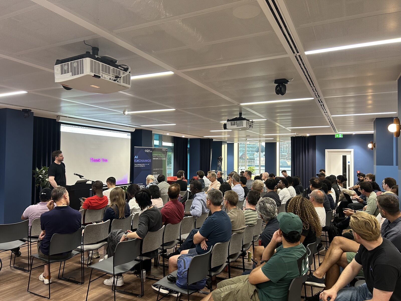 A speaker closing their talk in front of a “Thank You” slide, with the AI Exchange banner beside the stage and the audience seated in rows.